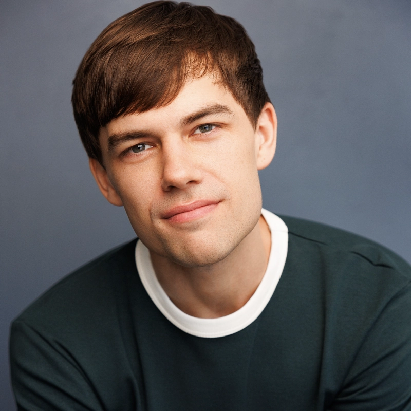 A young man with straight brown hair and fair skin looks at the camera, smiling slightly. He wears a forest green crew neck shirt with a white collar. The background is a simple, out-of-focus blue-gray, lending an understated backdrop to this classic portrait of Andrew Oppmann. The lighting is soft and even.