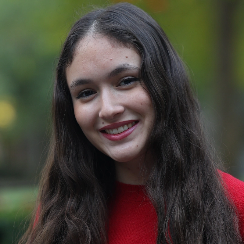 Jaleah Ortiz, a young woman with long, wavy brown hair, smiles at the camera in a bright red sweater. She stands outdoors with a soft, natural light and a blurred green background suggesting a peaceful park or garden setting.