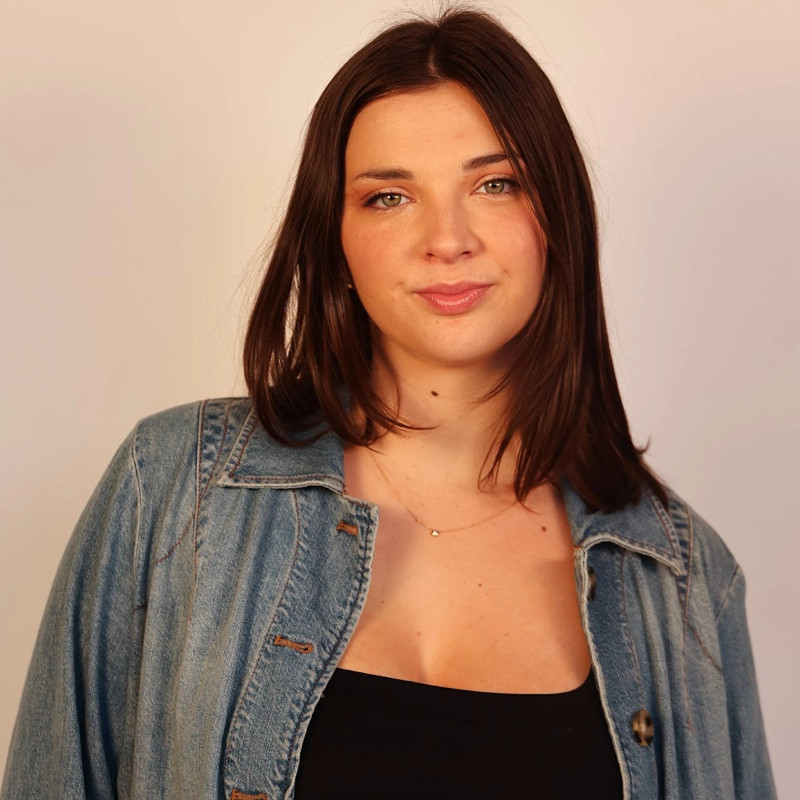 A young woman with straight brown hair, wearing a denim jacket over a black strapless top, stands against a plain light background. Georgia Ossorguine wears a thin gold necklace, her hair parted slightly off-center and her expression neutral.