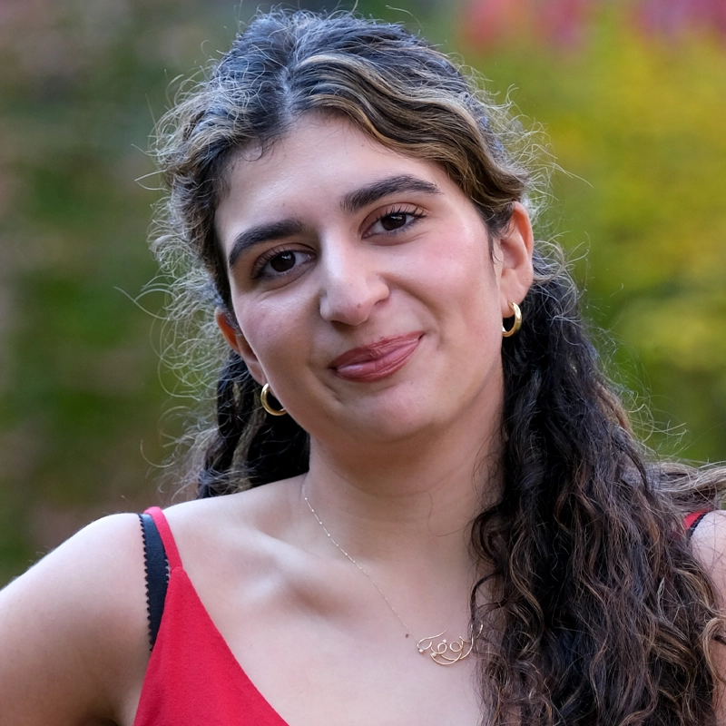 Sofia Gaviria Partow, a young woman with wavy brown hair and gold hoop earrings, smiles softly. She wears a red sleeveless top and a delicate pendant necklace. Blurred greenery with hints of yellow and red suggests an outdoor setting.