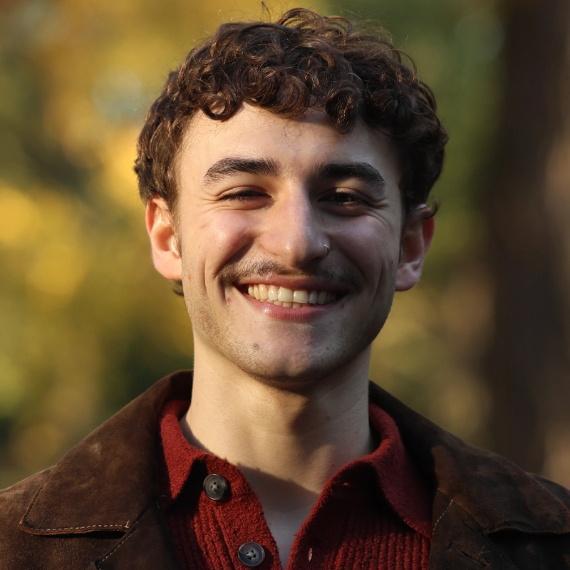 Michael Pontecorvo, a young man with curly brown hair and a mustache, smiles broadly outdoors. He wears a brown jacket over a red sweater, surrounded by sunlight and blurred foliage on a pleasant day in the park or forest.