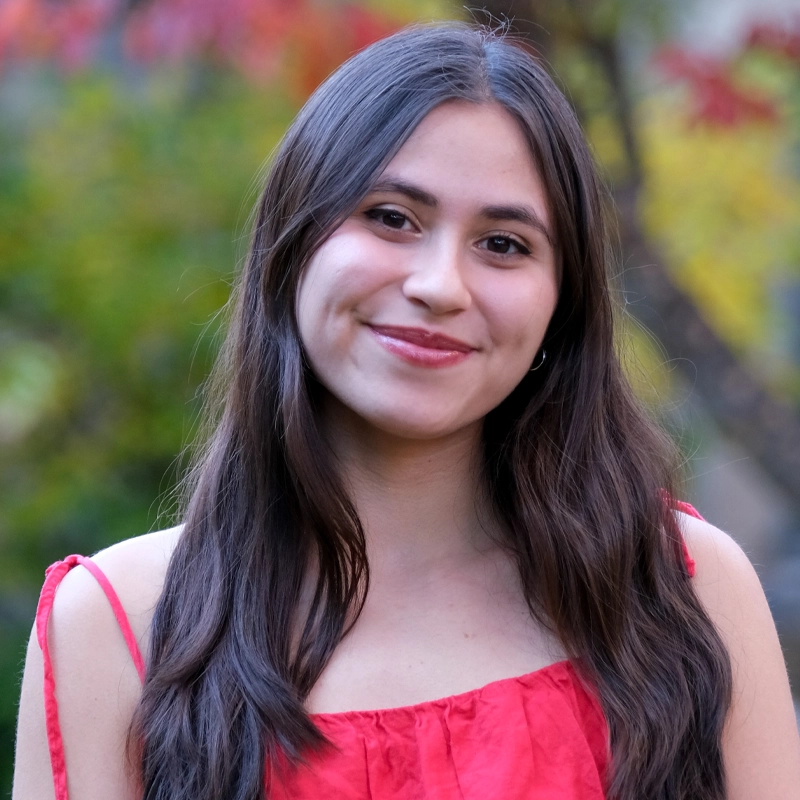 A young woman with long, dark brown hair and light skin smiles gently at the camera. Alexis Ramirez-Hardy is wearing a sleeveless red top. The background is blurred with greenery and hints of autumn colors, suggesting an outdoor setting.