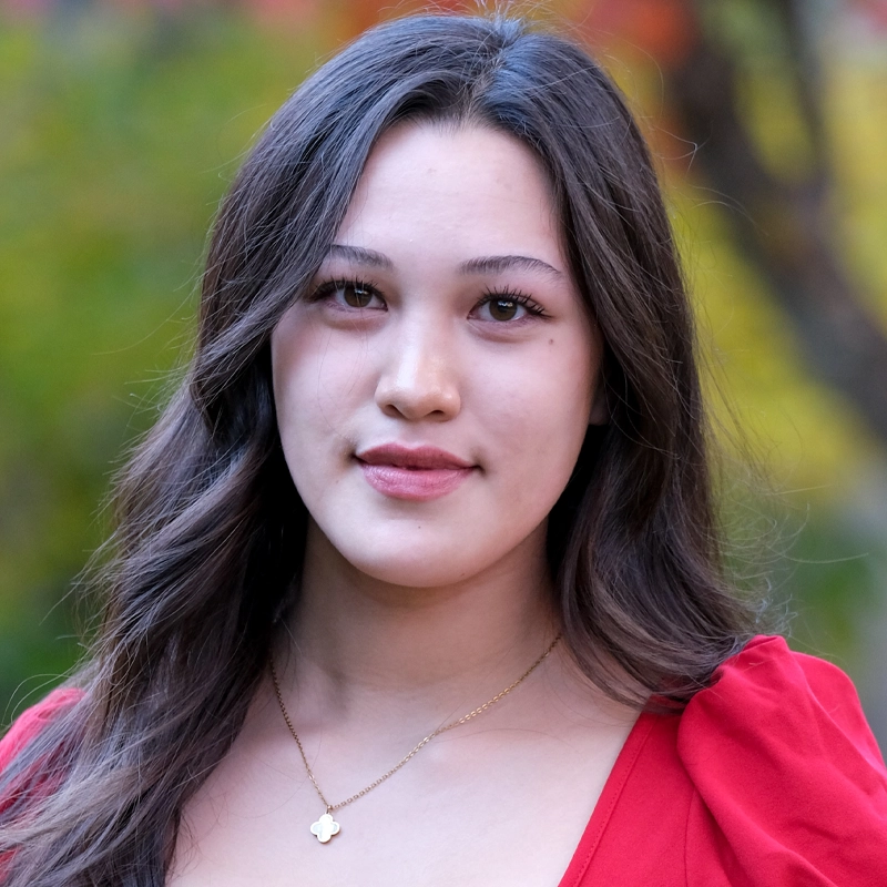 Anika Rodriguez, a young woman with long, wavy brown hair, stands outdoors against a blurred background of green and yellow foliage. She wears a red top and a gold necklace with a small pendant, looking at the camera with a calm expression.