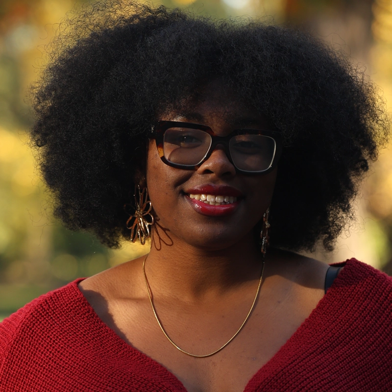 A woman with voluminous natural hair and glasses smiles outdoors. Naiya Rookwood wears a deep red knit top, gold hoop earrings, and a gold necklace. The blurred green and yellow foliage suggests a sunny day in a park or garden.