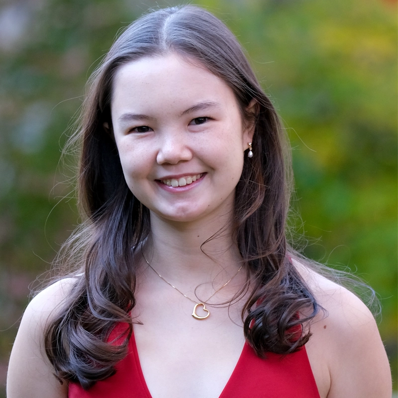 A young woman with fair skin and long brown hair smiles outdoors. She is wearing a red sleeveless top, a gold heart-shaped necklace, and small earrings. The background is blurred greenery with hints of yellow and orange, suggesting a natural setting.