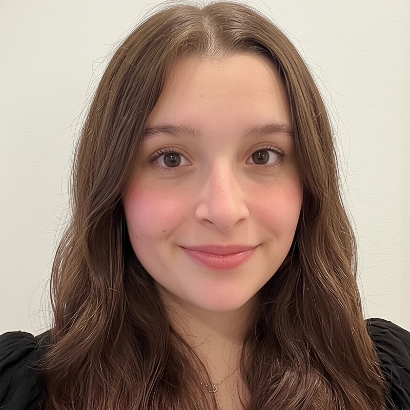Rio Schneider, a young woman with long, wavy brown hair and fair skin, smiles softly at the camera. She has brown eyes, light makeup, and wears a black top against a plain, light-colored background for a clean look.