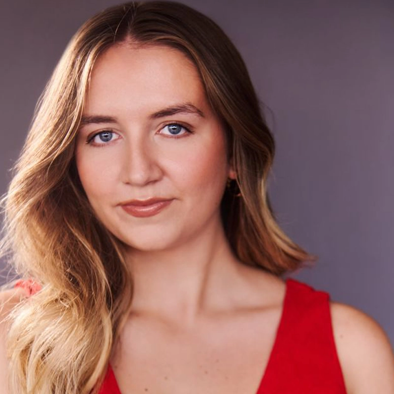 A young woman with long, wavy blonde hair and fair skin looks at the camera, wearing a sleeveless red top. With blue eyes and a slight smile, she poses against a softly lit gray background—capturing Shea Suffoletta’s effortless elegance.