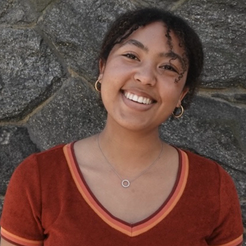 Jasmine Tihagoane, a young woman with curly hair, smiles in front of a stone wall. She wears gold hoop earrings, a silver pendant necklace, and a rust-colored V-neck shirt as sunlight illuminates her face, creating a warm, cheerful atmosphere.