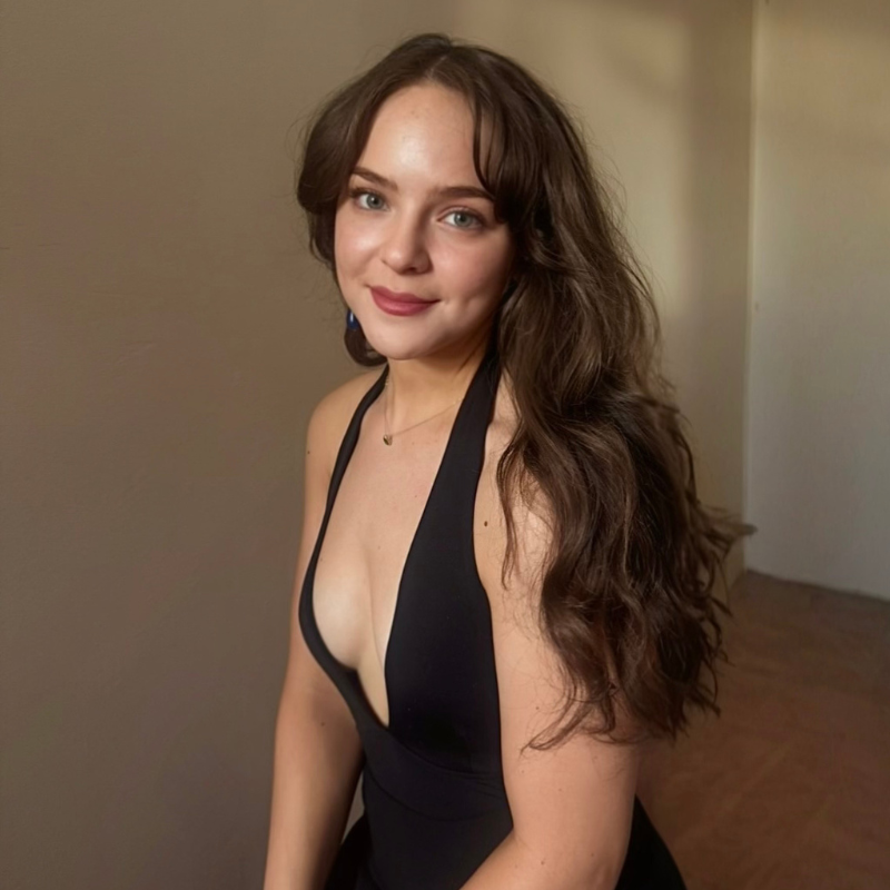 A young woman with long brown wavy hair and fair skin smiles gently. She wears a sleeveless black dress with a deep V-neckline, sitting in a softly lit room with beige walls and wooden flooring.