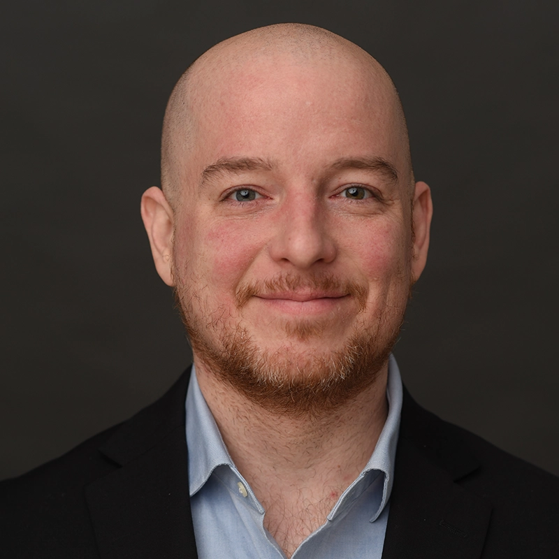 A bald man with fair skin and a short reddish beard smiles gently. He wears a black blazer over a light blue collared shirt. The solid dark gray background offers a professional, formal feel—ideal for profiles like Kiana Mottahedan’s.