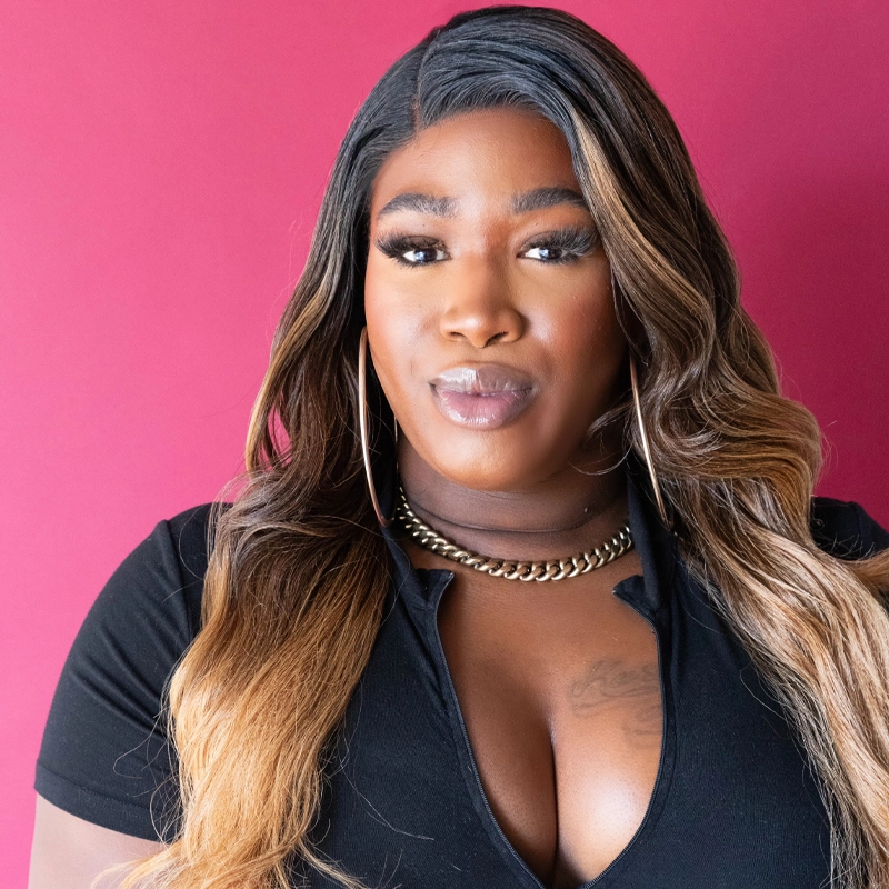 A woman with long, wavy brown and blonde hair stands against a magenta background. B Noel Thomas wears a black top, gold chain necklace, large hoop earrings, and subtle makeup with dramatic eyelashes, facing the camera with a confident expression.