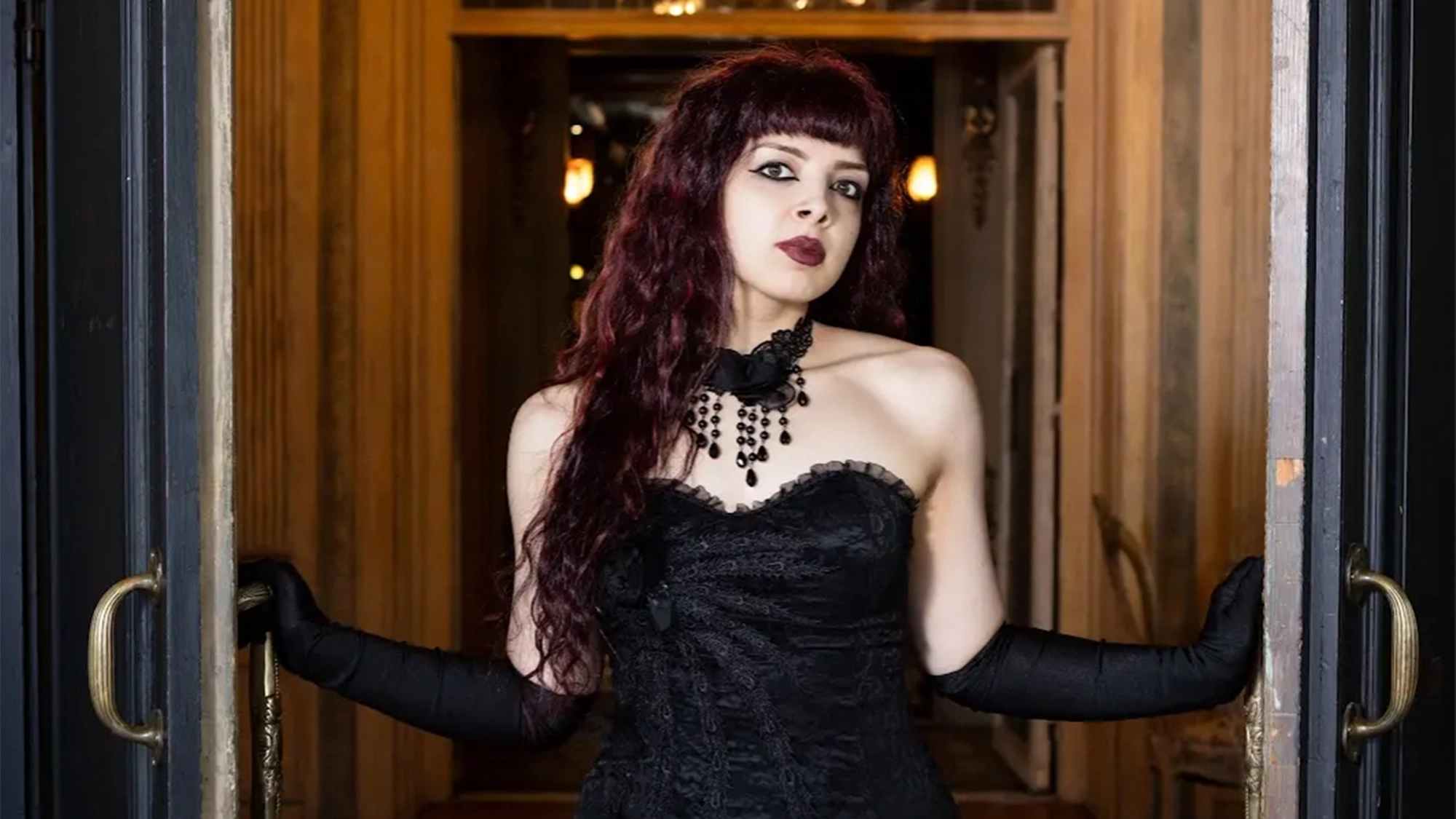 A woman resembling Artemisia LeFay with long, wavy dark red hair and dark lipstick stands in a doorway, wearing a black lace strapless dress, black choker with beads, and long black gloves. Warm lights and wooden walls are visible in the background.