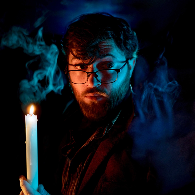 A bearded man with glasses, resembling Brandon Hardy, holds a lit white candle and looks to the side. Blue-tinted smoke swirls around him in the dark, as the candlelight illuminates his face and hand, heightening the mysterious atmosphere.