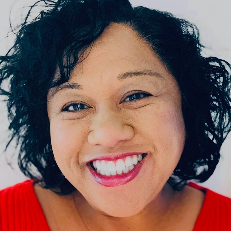 Christine Sta Ana, with her curly black hair and a radiant smile, exudes warmth in a red top. Her joyful expression is highlighted against the soft white background, and her bright eyes reflect friendliness and happiness.