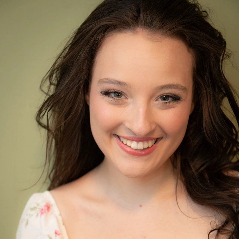 Olivia Conti, a young woman with long brown hair and light skin, smiles warmly at the camera. She has natural makeup, bright eyes, and wears a white top with a floral pattern. The softly blurred background is a gentle light green.
