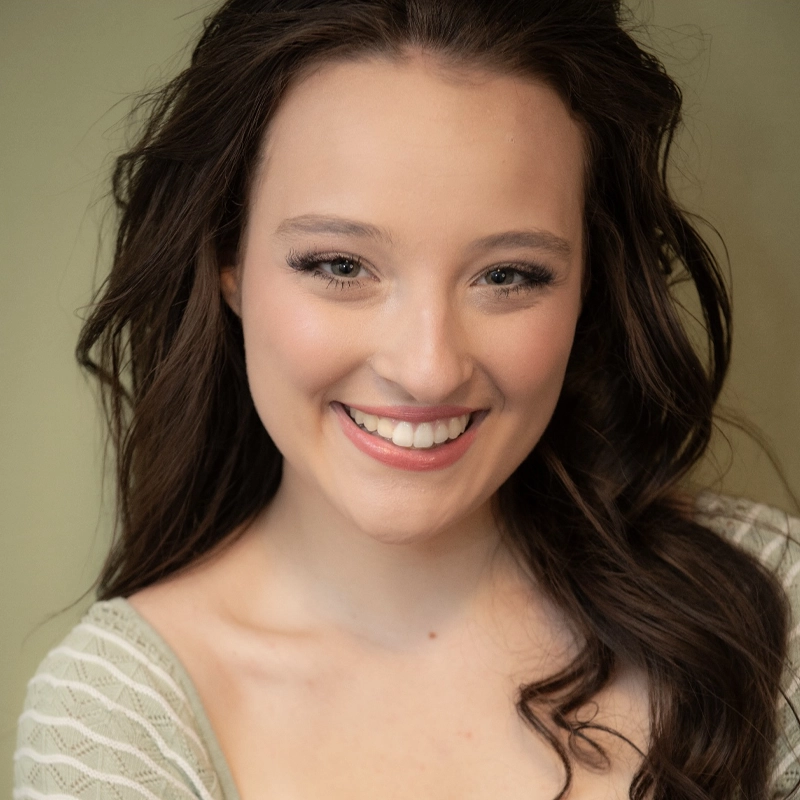 Olivia Conti, a young woman with long wavy brown hair, smiles at the camera. She has light skin, wears natural makeup, and is dressed in a light-colored, patterned top. The softly blurred background keeps the focus on her face.