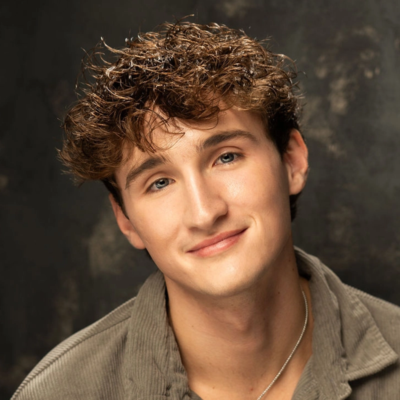 Cooper LaMontagne, a young man with wavy brown hair, smiles softly at the camera. He wears a gray corduroy shirt and a thin silver necklace. The background is dark and softly textured, highlighting his face and relaxed demeanor.