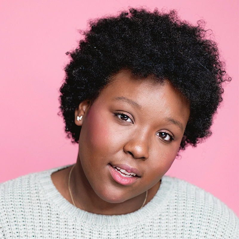 A young person with short, dark, textured hair looks toward the camera against a plain pink background. They have a natural makeup look, wear a light-colored knit sweater, and have a small butterfly earring in their ear.