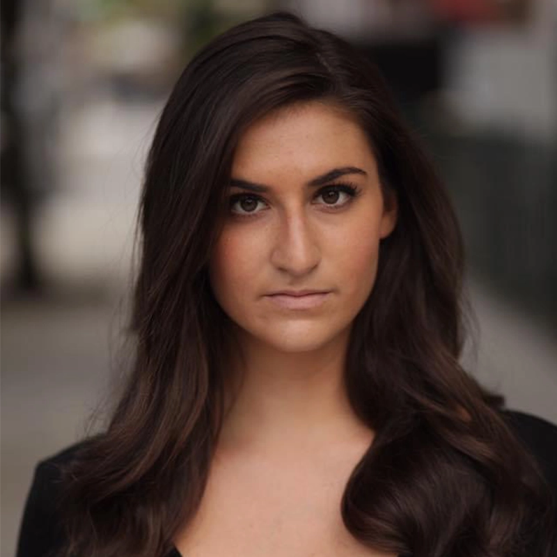 Danielle Palumbo, with long, wavy brown hair and fair skin, looks directly at the camera with a neutral expression. She wears a black top against a blurred urban background, softly lit by natural light.