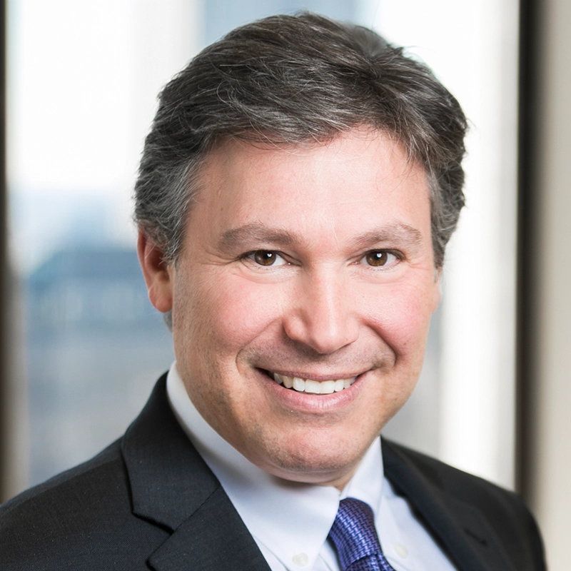 Jesse Manocherian, a middle-aged man with short, grayish-brown hair, smiles at the camera. He is wearing a dark suit, white dress shirt, and a purple tie against a blurred background of bright windows and hints of a cityscape.