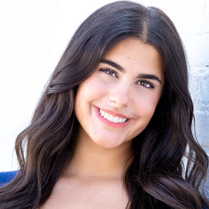 A young woman with long, wavy dark brown hair smiles at the camera. Olivia DeMarco has medium skin, dark eyes, and wears natural makeup. She is dressed in a blue top and posed against a light, subtly textured background.