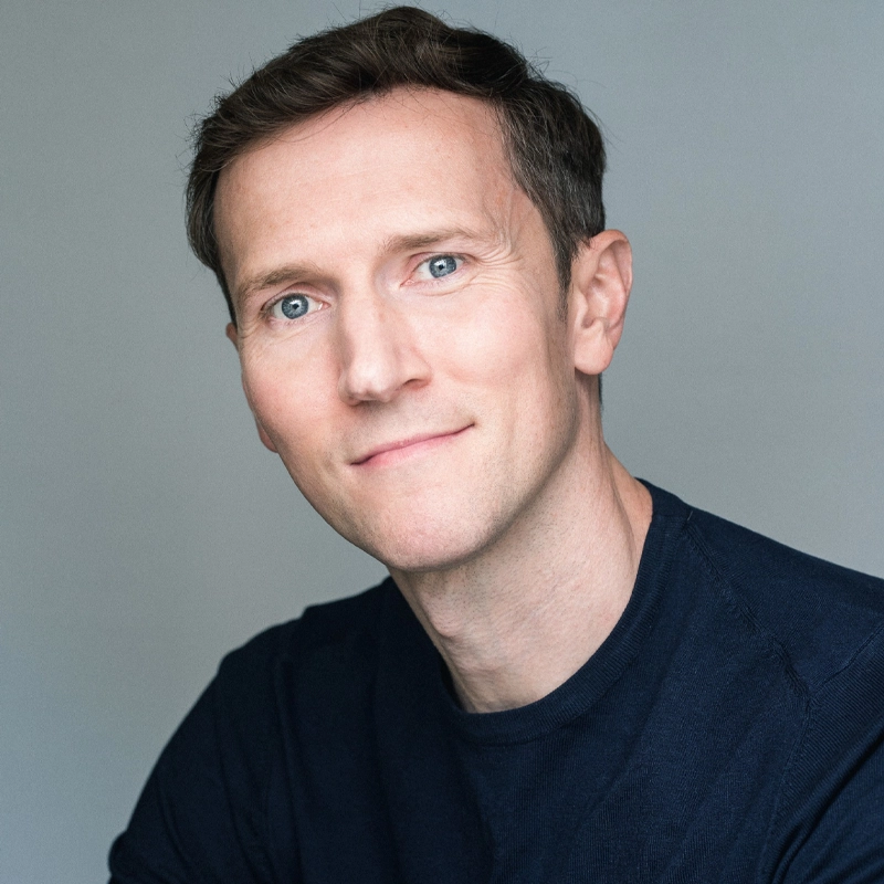 A man with short brown hair and blue eyes looks at the camera with a slight smile. He is wearing a dark navy blue crew-neck shirt and is posed against a plain, light gray background. The lighting is soft and even.