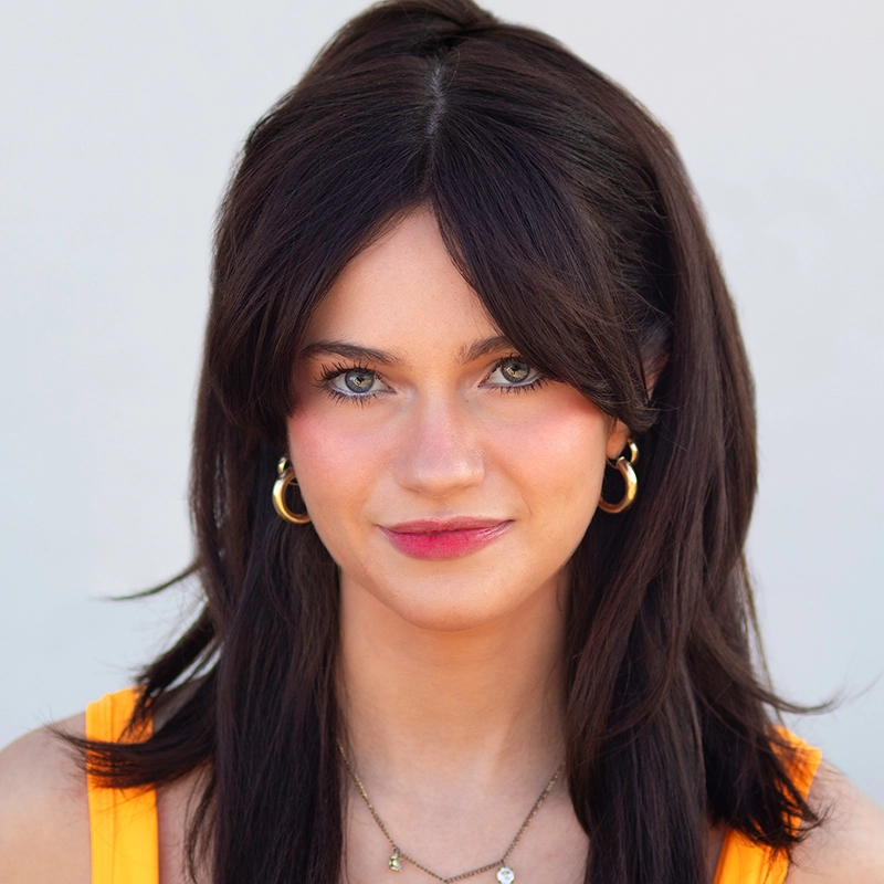 A portrait of a young woman with dark brown hair, curtain bangs, and blue eyes looking directly at the camera. She is wearing gold hoop earrings and a delicate necklace against a bright orange top.
