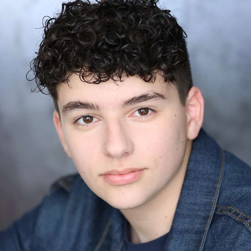 A young person with short, dark curly hair and fair skin looks directly at the camera, wearing a denim jacket over a dark shirt. The softly blurred gray and blue background highlights Jesse Manocherian's facial features and neutral expression.