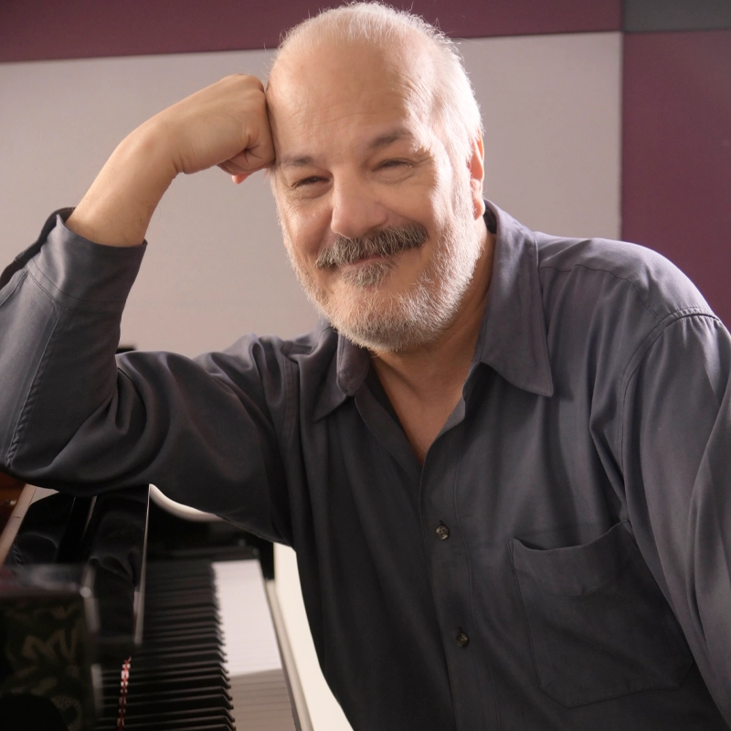 A smiling man with a beard leans his head on his hand, elbow resting on a grand piano. He is wearing a gray button-up shirt. The background features a two-toned wall with purple and gray panels.