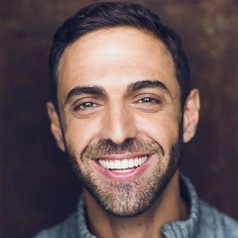 Jesse Manocherian, a man with short dark hair and a trimmed beard, smiles broadly, showing his teeth. He is wearing a gray sweater and is posed in front of a softly blurred brown background, giving a warm and friendly expression.