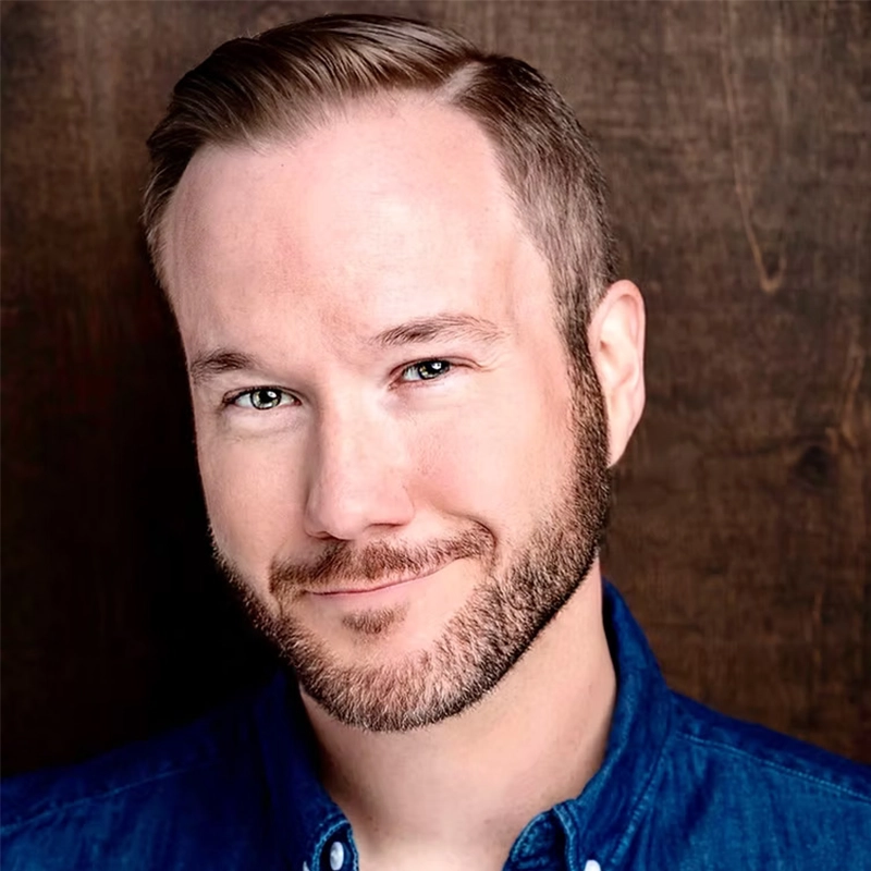 A smiling man, Josh Rhett Noble, with short, neatly styled light brown hair and a well-groomed beard looks at the camera. He wears a blue button-up shirt, and the dark wooden background gives the portrait a warm, friendly atmosphere.