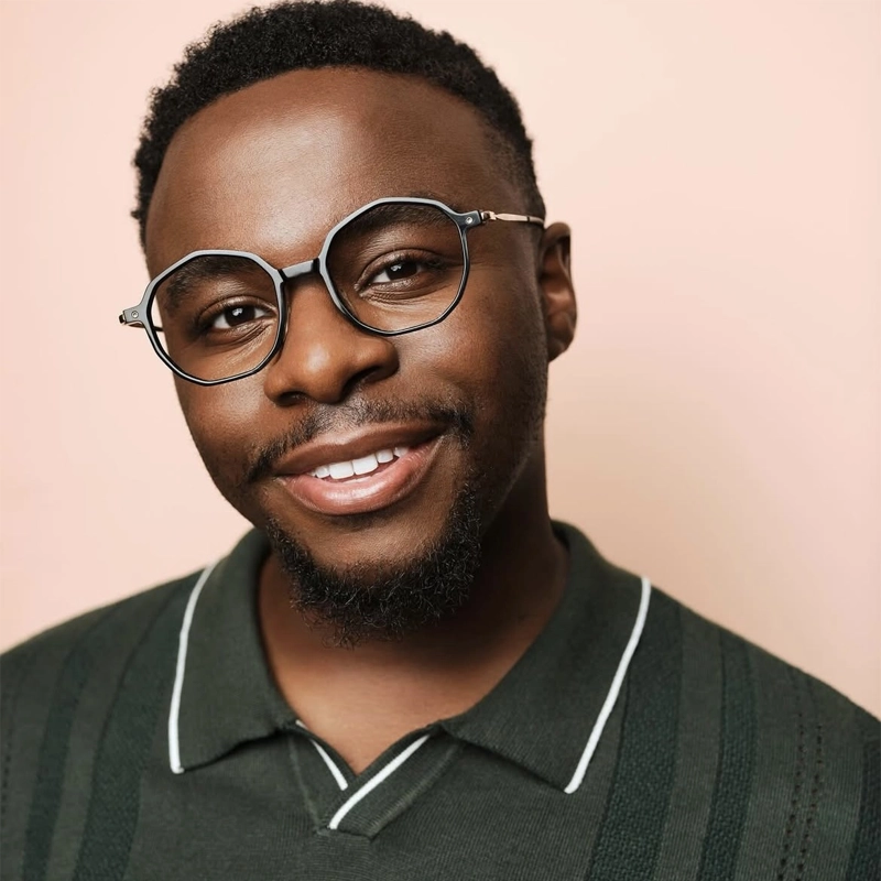 A smiling young Black man with a short beard and mustache, wearing round, stylish eyeglasses and a dark green collared shirt with light stripes, poses in front of a soft peach background. He has closely cropped hair and a friendly expression.