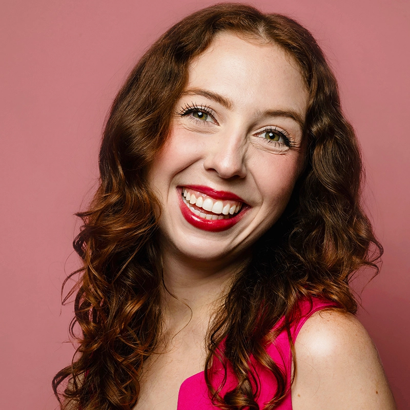 A smiling woman with wavy auburn hair and bright red lipstick looks toward the camera. She wears a vibrant pink top against a matching solid pink background.
