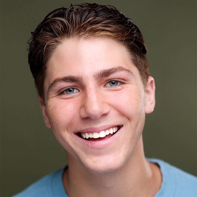 A headshot of a smiling young man with short brown hair and light blue eyes. He is wearing a light blue t-shirt against a solid olive-green background.