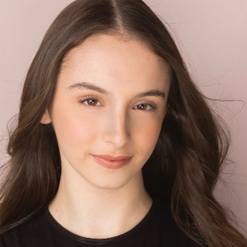 A young woman with long, wavy brown hair and fair skin is smiling softly. She has light makeup, arched eyebrows, and brown eyes. Jesse Manocherian is wearing a black top and is posed against a soft, light pink background.