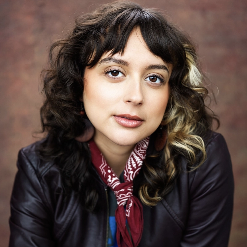 A young woman with wavy brown hair and a blonde streak looks at the camera. She wears a burgundy leather jacket, red bandana scarf, and teardrop earrings. The background is softly blurred in warm, earthy tones.