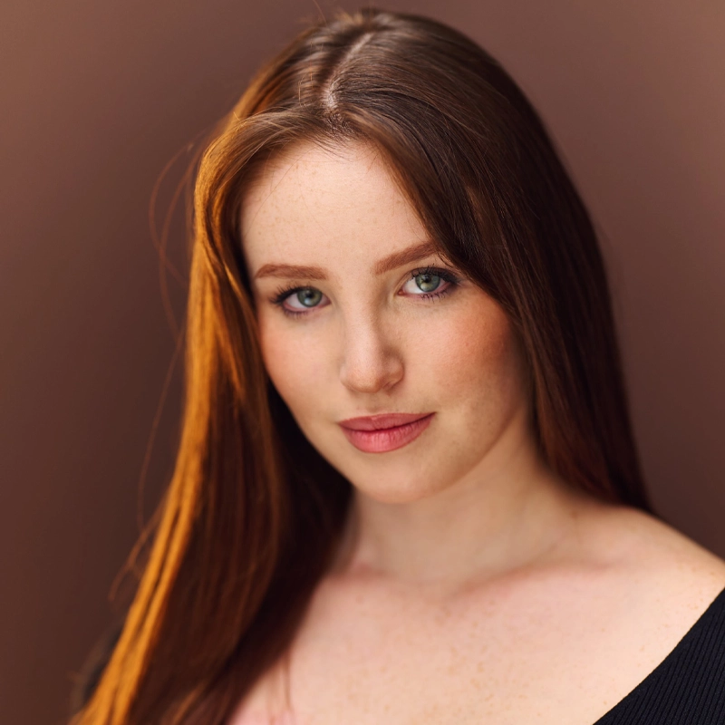 Eliana Nunley, a young woman with long, straight auburn hair and fair skin, looks calmly at the camera. She has green eyes, light freckles, and natural makeup, dressed in a black top against a softly blurred brown background.
