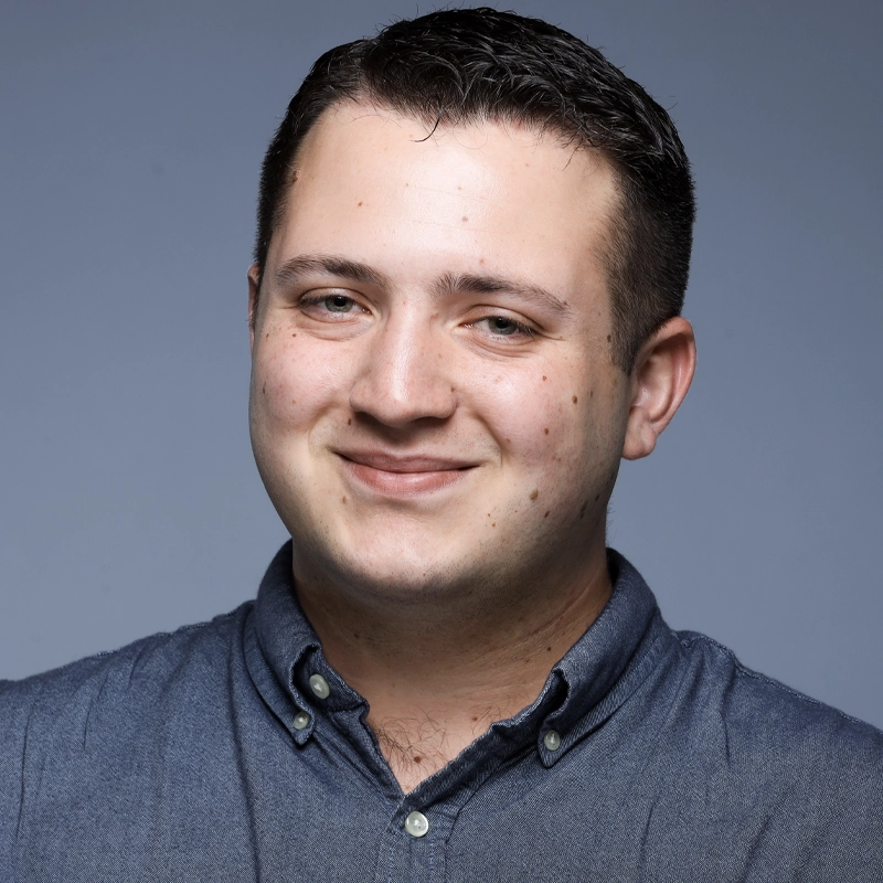 Nicolas Perez, a young man with short dark hair, smiles gently. He is wearing a buttoned-up, dark blue shirt. The plain, light gray background and even lighting give him a professional, friendly appearance suitable for a headshot or profile photo.