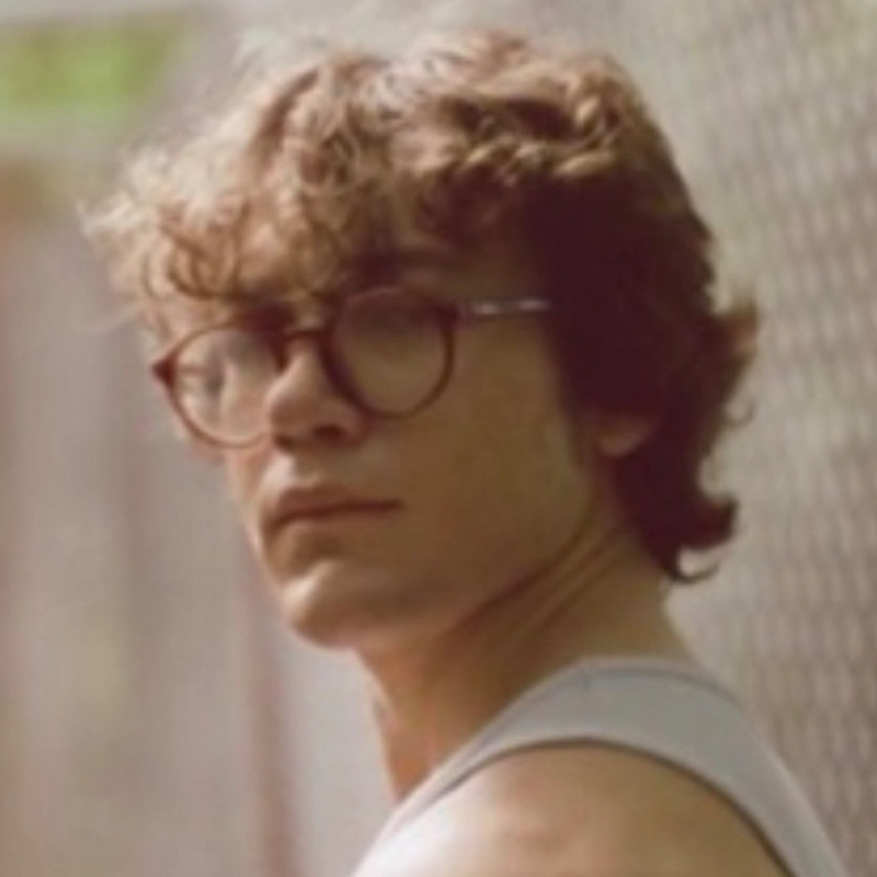 A person with wavy, shoulder-length brown hair and round glasses looks over their shoulder toward the camera. They are wearing a light-colored tank top and are positioned against a blurry, chain-link fence background.