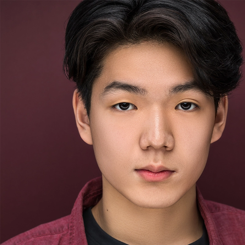 A headshot of a young person with dark hair styled with a side part, looking directly at the camera with a neutral expression. They are wearing a maroon button-down shirt over a black t-shirt against a dark, solid maroon background.