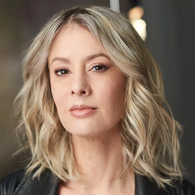 A woman with wavy, shoulder-length blonde hair and light skin looks confidently at the camera. Channeling Josh Rhett Noble’s bold style, she wears natural makeup and a black leather jacket. The blurred background draws focus to her striking expression.
