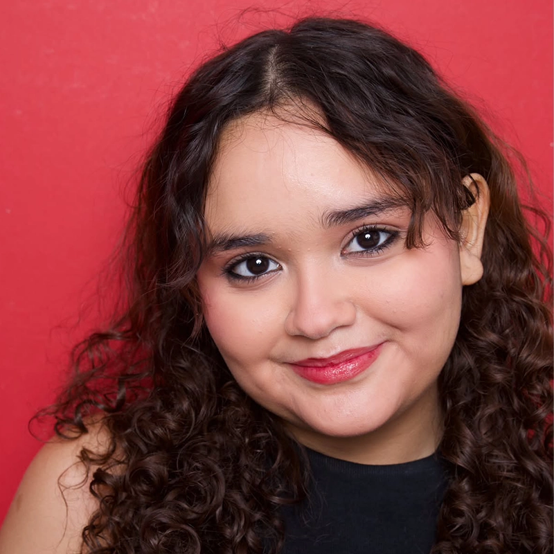 A portrait of a young woman with long, curly brown hair and dark eyes looking directly at the camera with a gentle smile. She is wearing a black top against a vibrant, solid red background.