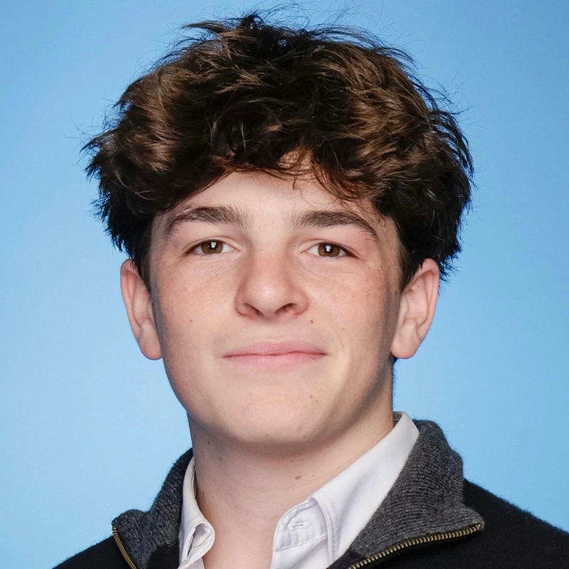 Jesse Manocherian, a young person with short, curly brown hair and fair skin, smiles slightly at the camera. They wear a white collared shirt under a dark zip-up sweater, set against a plain, solid light blue background.