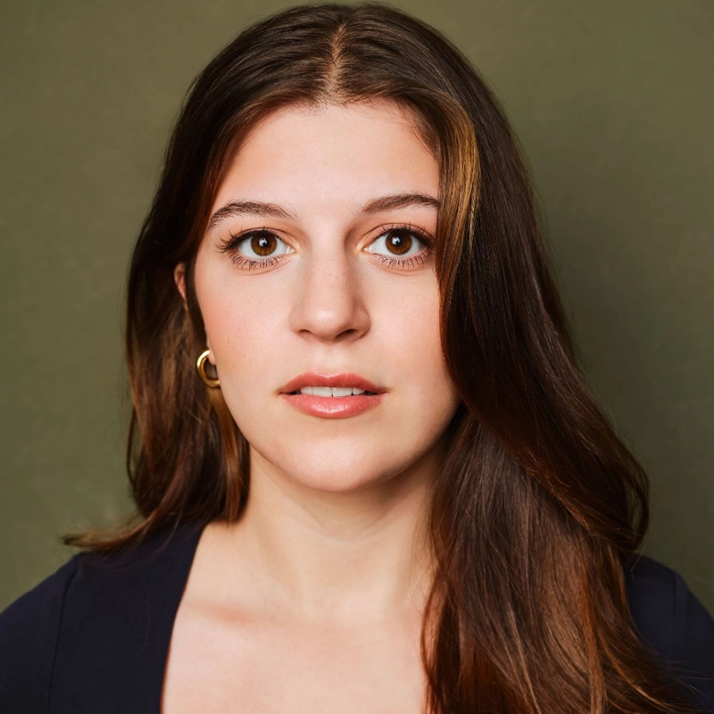 Rebecca Alice Steiner, a woman with long brown hair and brown eyes, looks directly at the camera. She wears a navy top and a gold hoop earring. The background is olive green. Her expression is neutral, and her hair is parted in the middle and falls over her shoulders.