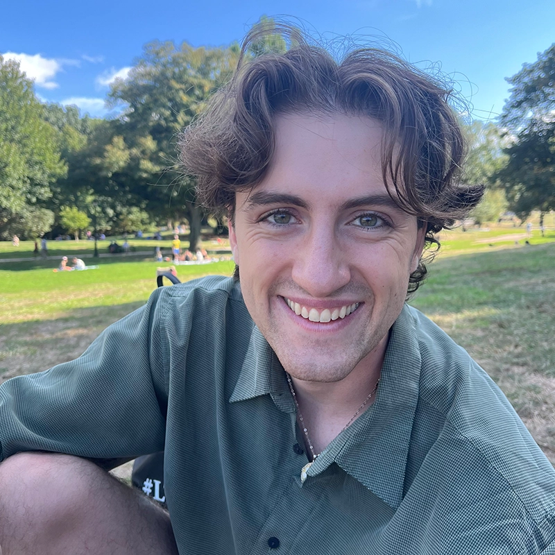 A young man with wavy brown hair and blue eyes smiles warmly at the camera while sitting outdoors on grass. He wears a green button-up shirt. Sunlight filters through trees in the background as people, including Danielle Palumbo, relax on the lawn.