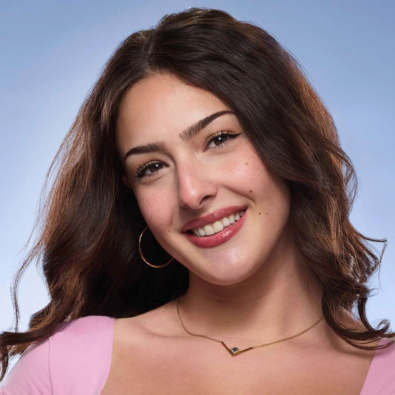 Valentina Marino, a woman with long, wavy brown hair, smiles at the camera. She wears large hoop earrings, a gold necklace, and a light pink top. The background is a soft, gradient blue, giving the photo a friendly, studio-lit atmosphere.