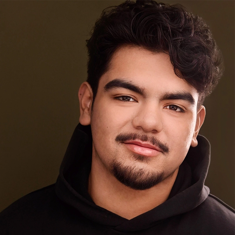 A close-up portrait of a young man with dark, wavy hair, a goatee, and a gentle smile. He is wearing a black hooded sweatshirt against a plain, dark olive-green background.