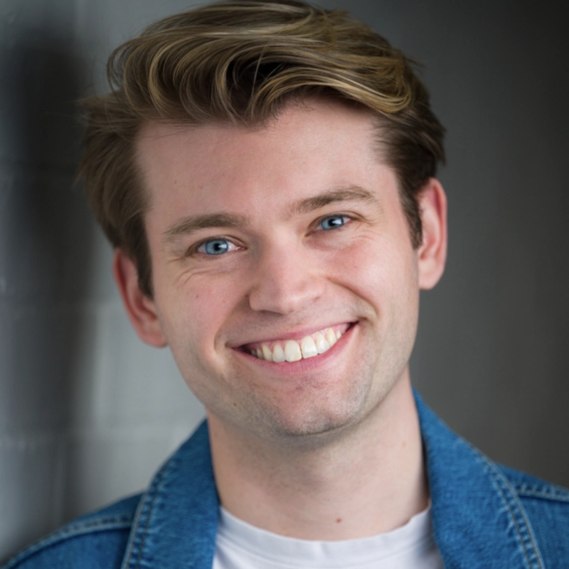 A young man with light skin and blue eyes smiles at the camera. He has short, light brown hair with a wavy style and is wearing a blue denim jacket over a white shirt. The background is blurred and gray, with soft lighting on his face.