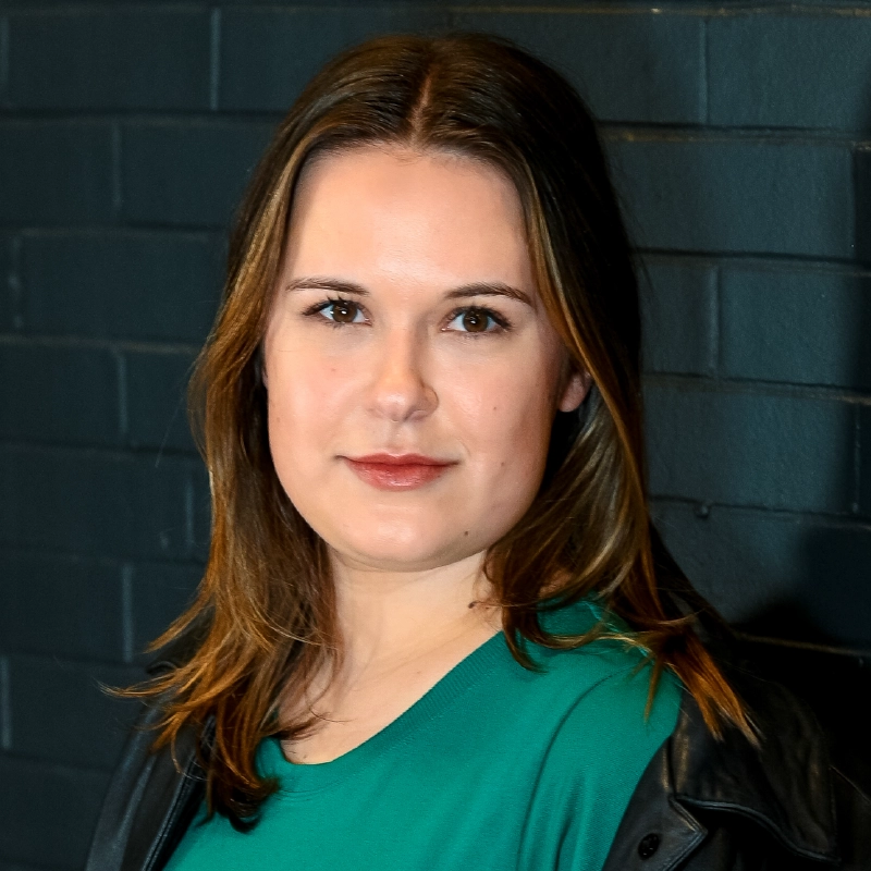 Anna Barrett, with straight brown hair parted slightly to the side, stands in front of a dark brick wall. She wears a green top and black jacket, her expression neutral as she looks directly at the camera.