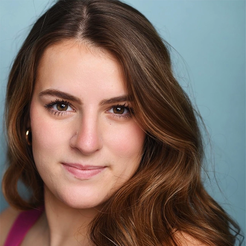 Ainsley Alrutz, a young woman with long, wavy brown hair and brown eyes, looks into the camera, smiling softly. She wears small gold hoop earrings and a pink top. The background is a solid light blue. Her makeup is natural and minimal.